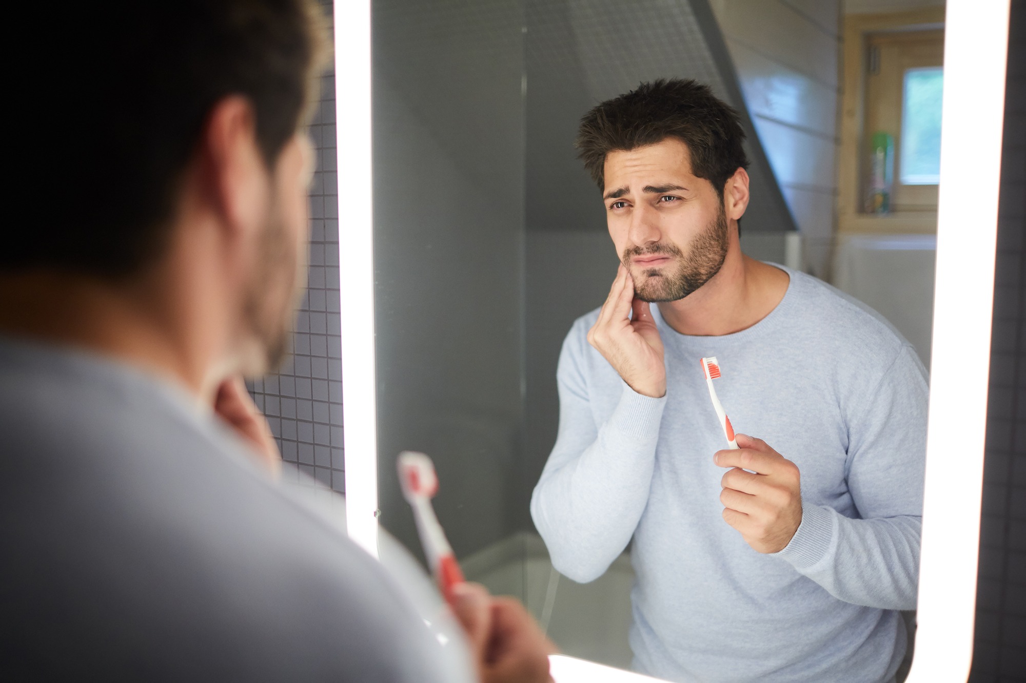 Man standing in the bathroom, looking in the mirror, showing a bad or decayed tooth